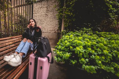 Şık ve genç gezgin Latin görünümlü esmer ve pembe valizli şehir parkını gezerken telefon kullanıyor.