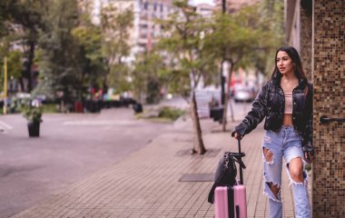 Şık seyahat: canlı Latin esmer moda görünümlü ve şehir merkezinde gezen pembe valizli.