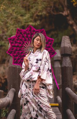 Tradition and beauty: japanese blonde woman in white silk kimono with flowers design and pink lace umbrella at wooden bridge in serene japanese park clipart