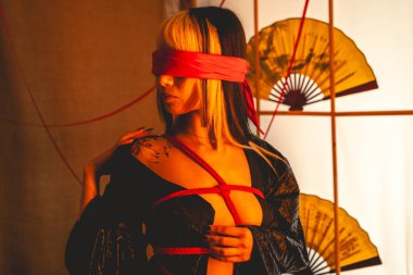 Sarı saçlı, siyah kimonolu, külotlu, kırmızı şii kravatlı, kırmızı gözlü, sıcak bir Japon odasında kağıt yelpaze tutan seksi ve güzel bir genç kadın.