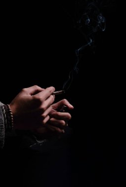 A vertical shot of unrecognizable hands holding a tobacco lit cigarette in a black background studio session. Smoke is coming out of the cigarette. Copy space for titles. Concept: addiction