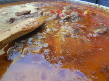 Et, sebze ve tencerede zengin et suyu ile kaynayan sığır eti yahnisinin yakın çekimi. Bir ahşap kaşık sıvının içinde duruyor, kaynayan pişirme işlemini gösteriyor. Kır manzarası geleneksel konforlu yiyecekleri vurguluyor.