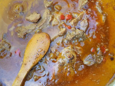 Et, sebze ve tencerede zengin et suyu ile kaynayan sığır eti yahnisinin yakın çekimi. Bir ahşap kaşık sıvının içinde duruyor, kaynayan pişirme işlemini gösteriyor. Kır manzarası geleneksel konforlu yiyecekleri vurguluyor.