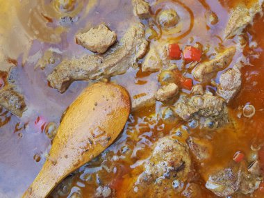 Et, sebze ve tencerede zengin et suyu ile kaynayan sığır eti yahnisinin yakın çekimi. Bir ahşap kaşık sıvının içinde duruyor, kaynayan pişirme işlemini gösteriyor. Kır manzarası geleneksel konforlu yiyecekleri vurguluyor.