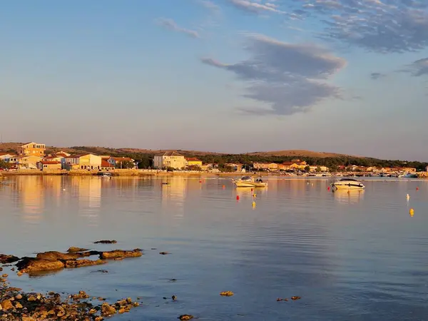Gün batımında, sakin suların sıcacık altın ışığı yansıttığı pitoresk bir sahil köyü. Tekneler limana demirlemiş ve kıyıya renkli evler dizilmiştir. Huzurlu sahne deniz kenarında yaşamanın güzelliğini yakalar..
