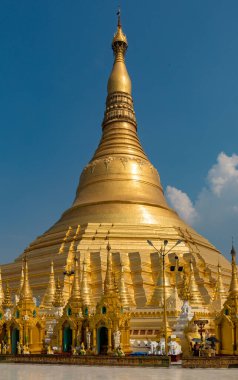 Açık mavi gökyüzünün altında süslenmiş, ön planda yürüyen insanların olduğu devasa bir altın tapınak. Myanmar.