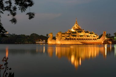 Aydınlatılmış Karaweik Sarayı 'nın göz kamaştırıcı bir gece manzarası, Kandawgyi Gölü' nde yüzen altın bir mavna, ışıkları sakin suyu yansıtıyor..