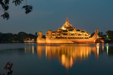 Aydınlatılmış Karaweik Sarayı 'nın göz kamaştırıcı bir gece manzarası, Kandawgyi Gölü' nde yüzen altın bir mavna, ışıkları sakin suyu yansıtıyor..