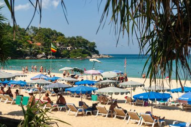 Kumun üzerinde güneşli şezlonglar ve şemsiyelerle dolu, turistlerin yemyeşil yapraklarla çerçevelenmiş gevşeyip yüzdüğü canlı bir tropikal plaj sahnesi. Tayland, Phuket