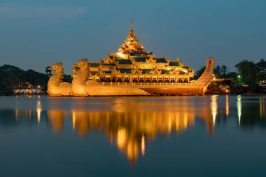 Aydınlatılmış Karaweik Sarayı 'nın göz kamaştırıcı bir gece manzarası, Kandawgyi Gölü' nde yüzen altın bir mavna, ışıkları sakin suyu yansıtıyor..