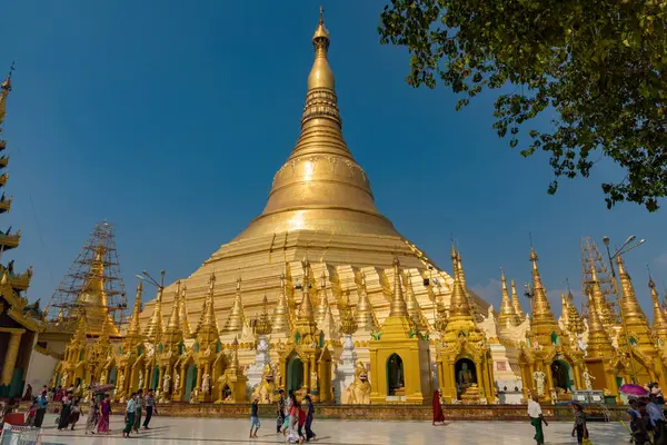 Açık mavi gökyüzünün altında süslenmiş, ön planda yürüyen insanların olduğu devasa bir altın tapınak. Myanmar.