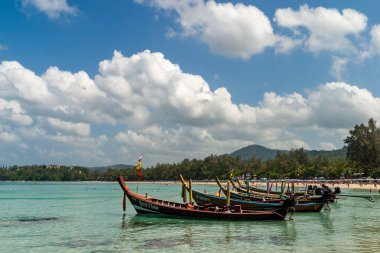 Tayland, Phuket. Geleneksel Tayland uzun kuyruklu tekneleri, dramatik bir gökyüzünün altında kumlu kıyıları ve palmiye ağaçları olan tropik bir plajın sığ ve berrak sularına demirlemişti..