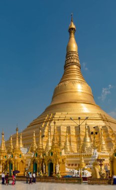 Açık mavi gökyüzünün altında süslenmiş, ön planda yürüyen insanların olduğu devasa bir altın tapınak. Myanmar.