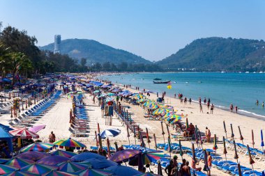 Renkli şemsiyeleri, şezlongları ve açık mavi gökyüzü altında turistleri olan kalabalık bir plaj sahnesi. Tayland Phuket