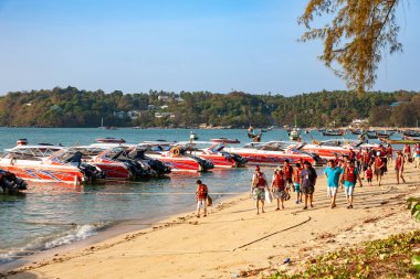 Can yelekli turistler kumlu bir sahil boyunca kırmızı sürat teknesi filosuna doğru yürüyorlar.