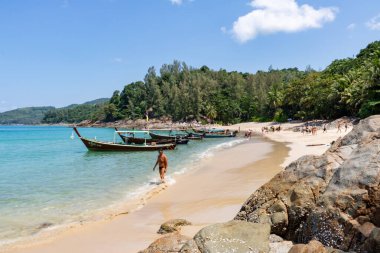Adam Phuket 'te güneşli bir plajda uzun kuyruklu teknelerle yürüyor.