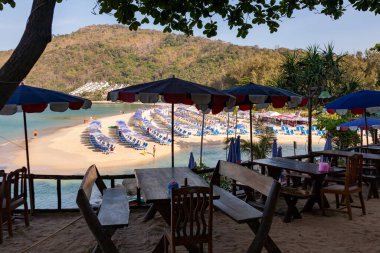 Sahil kenarındaki restoran masaları ve kumsal manzaralı şemsiyeler, kanolar ve tropikal yamaçlar. Tayland Phuket