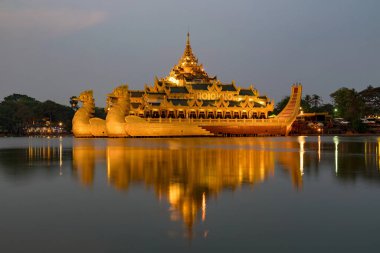Aydınlatılmış Karaweik Sarayı 'nın göz kamaştırıcı bir gece manzarası, Kandawgyi Gölü' nde yüzen altın bir mavna, ışıkları sakin suyu yansıtıyor..