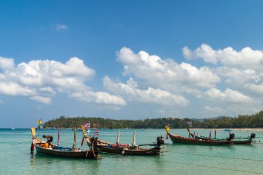 Tayland, Phuket. Güneşli bir günde, tropik bir plajın sığ ve berrak sularında demirlemiş geleneksel uzun kuyruklu tekneler grubu..