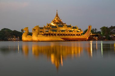 Aydınlatılmış Karaweik Sarayı 'nın göz kamaştırıcı bir gece manzarası, Kandawgyi Gölü' nde yüzen altın bir mavna, ışıkları sakin suyu yansıtıyor..