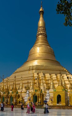 Açık mavi gökyüzünün altında süslenmiş, ön planda yürüyen insanların olduğu devasa bir altın tapınak. Myanmar.