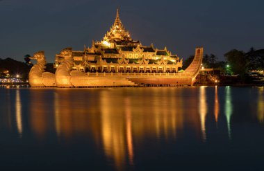 Aydınlatılmış Karaweik Sarayı 'nın göz kamaştırıcı bir gece manzarası, Kandawgyi Gölü' nde yüzen altın bir mavna, ışıkları sakin suyu yansıtıyor..