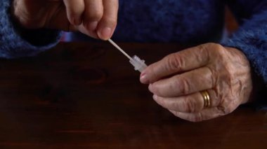 Kovid19 için antijen teçhizatıyla kendi kendine test yapan yaşlı bir kadının yakın çekimi. Kafkasyalı büyükanne Coronavirus enfeksiyonunu kontrol etmek için tampon damlatıcıya burun çubuğu takıyor..
