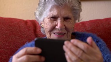 Kanepede otururken cep telefonu kullanan yaşlı bir kadın. Gülümseyen mutlu büyükannenin elinde akıllı telefon var. Yaşlı büyükanne telefon kullanıyor. Teknoloji aygıtı kavramı