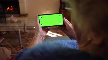 Evde yeşil ekranlı cep telefonu kullanan yaşlı bir kadın var. Elementleri eklemek için bir model aygıtı. Büyükannenin buruşuk elleri akıllı telefon tutuyor. Teknoloji kavramı