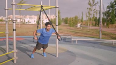 Caucasian man exercising outside in sunny park. Great TRX workout. Young handsome guy in sportswear doing exercising outdoors. Healthy lifestyle concept.