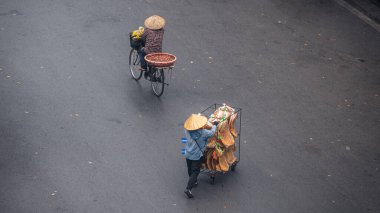 Bisikletli biri bisikletleriyle asfalt bir yol boyunca bisiklet sürerken, hava manzaralı sokak yiyecek satıcısı bir araba dolusu aperatifi itiyor. Her iki kişi de geleneksel Asya konik hasır şapkaları takıyor.