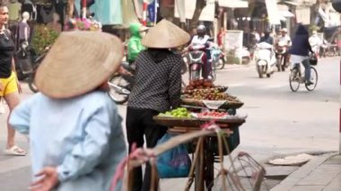 Vietnam, Hanoi-03 Nisan 2024: Geleneksel Asya konik hasır şapkası takmış Vietnamlı bir sokak satıcısının yavaş çekimi, bisikletini Hanoi 'de bir yol boyunca seyyar meyve tezgahı olarak kullanıyor..