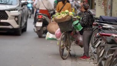 Vietnam, Hanoi-03 Nisan 2024: Geleneksel Asya konik hasır şapkası takmış Vietnamlı bir sokak satıcısı, bisikletini Hanoi yolu boyunca seyyar meyve tezgahı olarak kullanıyor.