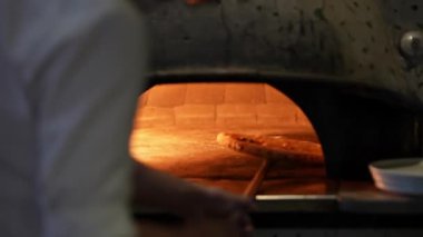 Profesyonel bir aşçı geleneksel tuğla fırınıyla çalışıyor. Isıtılmış taşın üzerine altın kahverengi kabuklu bir pizza kayıyor. Közlerin yoğun sıcaklığı pizzanın mükemmel pişmesini sağlar.