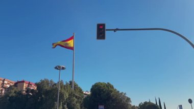 İspanya bayrağı açık mavi gökyüzüne karşı kırmızı bir trafik ışığının yanında gururla dalgalanıyor. Kentsel manzara ve vatanseverliği tek bir çerçevede yakalar.