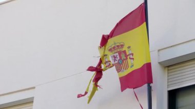 Bir direkten dalgalanan yıpranmış İspanyol bayrağının yavaş hareketi, kenarları yıpranmış, iplikleri gevşemiş ve kumaşı hafifçe yırtılmış. Kırmızı ve sarı renkler ve ulusal arma düz bir duvara karşı tezat