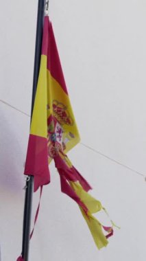 Dikey Ağır çekim, yıpranmış kenarlar, gevşek iplikler ve yırtık kumaş parçalarından oluşan yıpranmış bir İspanyol bayrağı. Kırmızı ve sarı renkler ve arma düz bir duvara karşı duruyor, direnç uyandırıyor..