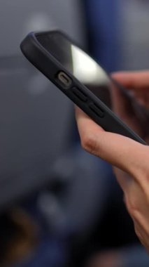 Yakın plan Asyalı kadın ellerinin uçakta akıllı telefon tutarken bulanık yolcular ve arka planda otururken çekilmiş dikey görüntüleri. Seyahat sırasında teknoloji kullanımı, gündelik ve modern atmosfer