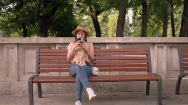 Şapkalı Asyalı bir kadın, huzurlu bir şehir parkında ahşap bir bankta, akıllı telefonuna odaklanmış oturuyor. Arka plandaki ağaçlar sakin bir ortam yaratır. Yumuşak gün ışığında doğa ve şehir yaşamını harmanlarlar.
