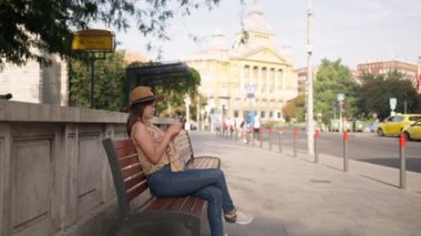 Şık şapkalı ve günlük kıyafetli Asyalı bir kadın, tarihi bir Avrupa binası olan Budapeşte 'de ahşap bir bankta akıllı telefonunu kullanarak oturuyor. Güneşli bir havada huzurlu bir an. Telefon kullanan biri.