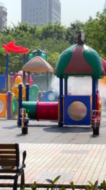 Dikey bir işçi renkli bir oyun alanını, güvenlik önlemlerini dezenfekte eder. Yüksek binaların arka planına sahip bir şehir parkında kamu sağlığı çalışmaları.