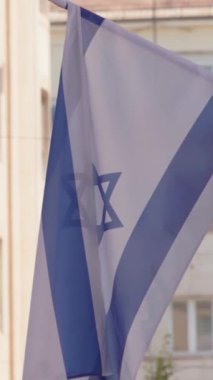 İsrail bayrağının dikey görüntüsü bir caddede güneşli bir binaya doğru dalgalanıyor. Kentsel ortamlardaki kültürel varlığı, uluslararası birliği ve mimari güzelliği vurguluyor.