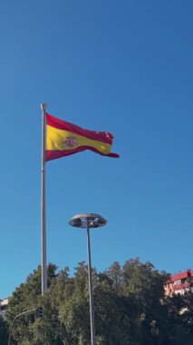 Dikey Ağır çekim yıpranmış kenarları, gevşek iplikleri ve yırtık kumaşı olan bir direkten eskimiş İspanyol bayrağı dalgaları. Kırmızı ve sarı renkler ve arma bir duvara karşı duruyor, dayanıklılık çağrıştırıyor.