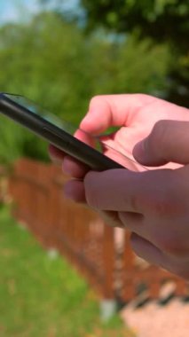 Güneşli bir bahçede akıllı telefonları kullanarak insan ellerine dikey çekim yapmak. Yeşil çevre ve arka plandaki ahşap çit huzurlu ve rahat bir atmosfer yaratıyor, modern telefon bağlantısı sağlıyor.