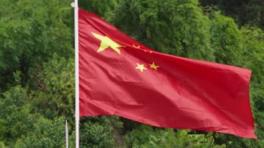 Bir Çin bayrağının yavaş hareketi ağaçların yemyeşil zeminine doğru dalgalanıyor, canlı kırmızı rengini ve altın yıldızlarını sergiliyor. Doğal ortam, ulusal bayrağın cesur renkleriyle çok güzel tezat oluşturuyor.