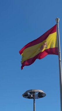 Bir direkten dalgalanan yıpranmış İspanyol bayrağının dikey yavaş hareketi, kenarları yıpranmış, iplikleri gevşemiş ve kumaşı hafifçe yırtılmış. Kırmızı ve sarı renkler ve ulusal arma bir duvara karşı