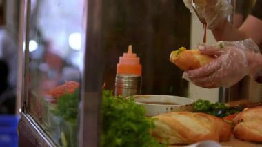 Hanoi, Vietnam-03 Nisan 2024: Şefin tipik Banh mi 'ye sos koymasının yavaş çekimi, et ve sebzeli sandviç. Lezzetli malzemelerle Fransız baharatlarını birleştirmiş bir baget..