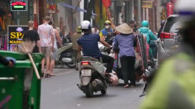 Hanoi, Vietnam-04 Nisan 2024: Geleneksel Asya konik hasır şapkası takan bir sokak satıcısının yavaş çekimi, turist hatıralarıyla dolu bir arabayı itiyor, bir trafik yolu şehrinde geziniyor.