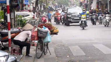 Hanoi, Vietnam-04 Nisan 2024: Geleneksel Asya konik hasır şapkası takmış Vietnamlı bir sokak satıcısı, bisikletini Hanoi, Vietnam 'da bir yol boyunca mobil meyve tezgahı olarak kullanıyor..