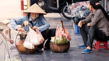 Hanoi, Vietnam-04 Nisan 2024: Geleneksel Asya konik hasır şapkasıyla süslenmiş kaldırımlarda yiyecek satan Vietnamlı bir kadın sokak satıcısı. Geleneksel mutfak